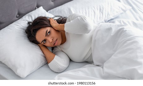 Portrait Of Irritated Young Black Woman Lying In Bed And Covering Ears With Hands, Hearing And Suffering From Too Loud Sound Or Noise. Lady Feeling Uncomfortable, Trying To And Can't Sleep