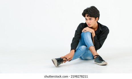 Portrait Isolated Cutout Studio Shot Of Asian Young LGBTQ Gay Glamour Handsome Male Model In Casual Black Shirt Jeans And Sneakers Sitting Crossed Legs Posing Gesturing On Floor On White Background.