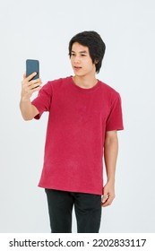 Portrait Isolated Cutout Studio Shot Of Asian Young Handsome Teenager Male Model In Street Style Outfit Standing Holding Smartphone In Hands Smiling Taking Selfie Photo On White Background.