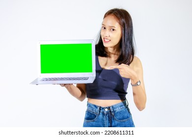 Portrait Isolated Studio Cutout Shot Of Asian Female Model Smiling Look At Camera Holding Pointing Green Blank Screen Laptop Computer Showing Product Promotion Advertisement On White Background.