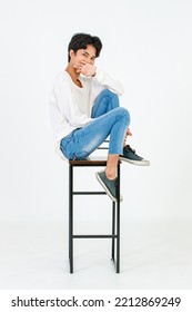 Portrait Isolated Studio Shot Asian Young LGBT Gay Happy Handsome Bisexual Homosexual Male Fashion Model In Casual Outfit Sitting Crossed Legs On Tall Chair Smiling Look At Camera On White Background.