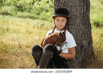 Portrait of a jockey girl in a helmet and a white T-shirt, who sits under a tree with a toy horse in her hands. Hobbyhorsing. - Powered by Shutterstock