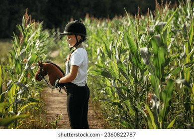 Portrait of a jockey girl in a helmet and a white t-shirt, who sits on a brown toy horse in a corn field. Hobbyhorsing. - Powered by Shutterstock