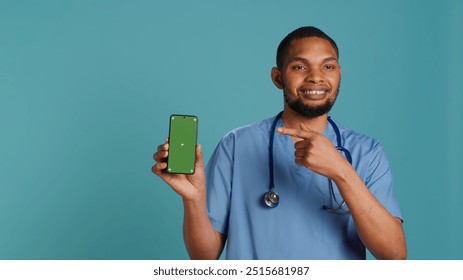 Portrait of jolly male nurse pointing finger towards green screen smartphone. Upbeat hospital employee holding chroma key phone, isolated over blue studio background, camera A - Powered by Shutterstock