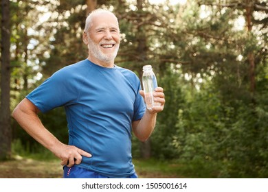 Portrait Of Joyful Active Caucasian Retired Man With Beard And Bold Head Holding Hand On His Waist And Drinking Fresh Water From Glass Bottle, Having Rest After Morning Physical Workout In Park