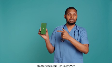 Portrait of joyful african american male nurse pointing finger towards green screen cellphone. Smiling healthcare professional holding mockup mobile phone, studio background, camera B - Powered by Shutterstock