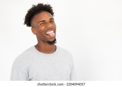 Portrait Of Joyful African American Man Sticking Out Tongue. Young Bearded Guy Wearing White Sweater Looking At Camera And Winking. Fun Concept