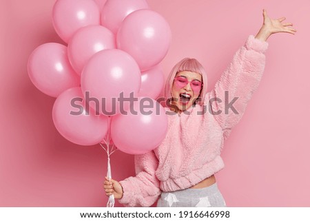 Portrait of joyful Asian woman with pink hair raises hand has fun exclaims happily finally passed all exams at university and has graduation party dressed in festive clothes holds bunch of balloons