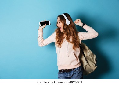 Portrait of a joyful attractive girl student with backpack listening to music with headphones while showing blank screen mobile phone and dancing isolated over blue background