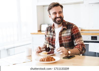 Portrait of joyful bearded man smiling and eating cake while having breakfast in kitchen at home - Powered by Shutterstock