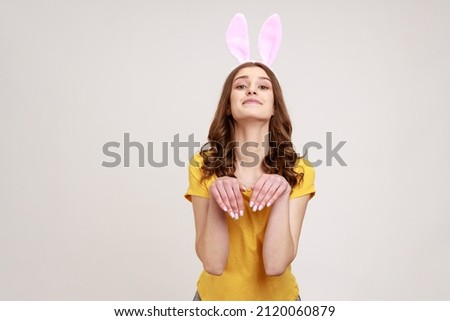 Portrait of joyful beautiful woman of young age in yellow T-shirt behaving childish humorous with bunny ears and hands, entertaining and fooling. Indoor studio shot isolated on gray background.