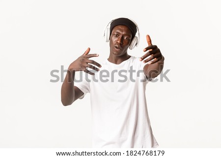 Portrait of joyful cool african-american guy, dancing and listening music in headphones, white background