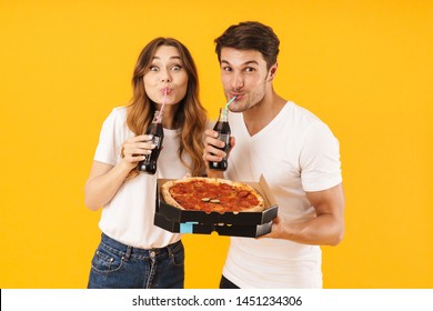 Portrait of joyful couple man and woman in basic t-shirts drinking soda beverage and holding pizza boxes isolated over yellow background