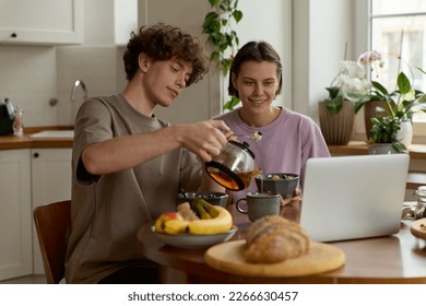 Portrait of joyful friends man and woman smiling pouring drinking tea together during breakfast in the kitchen while using wireless technology laptop watching films browsing on computer. Lifestyle.