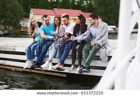 Portrait of joyful group of young people sitting on the edge of the pier, outdoors in nature. Friends enjoying a day on the lake