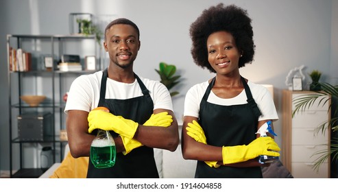 Portrait of joyful happy young African American workers standing in room in apartment in aprons looking at camera and smiling holding cleaning liquid spray, cleaning service concept, small business