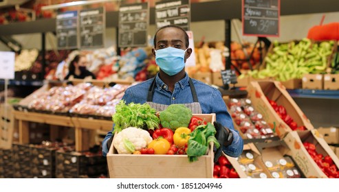 Portrait Of Joyful Male Food Store Manager In Mask And Gloves Standing In Supermarket And Holding Box With Fresh Vegetables, Looking At Camera And Smiling. African American Mam Working At Grocery.