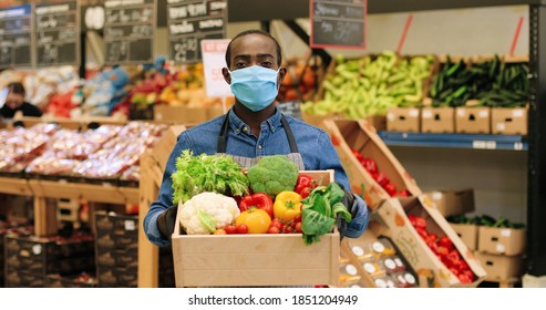 Portrait Of Joyful Male Food Store Manager In Mask And Gloves Standing In Supermarket And Holding Box With Fresh Vegetables, Looking At Camera And Smiling. African American Mam Working At Grocery.