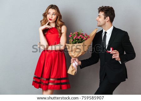 Portrait of a joyful man proposing to a unsatisfied girl with flowers and an engagement ring over gray wall background