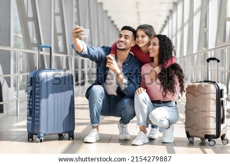 Portrait Of Joyful Middle Eastern Family Of Three Taking Selfie With Smartphone In Airport Terminal, Happy Arab Parents And Little Daughter Posing To Camera While Sitting Next To Suitcases