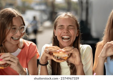 Portrait of joyful smiling teenage girls (14-15 years old) sitting at a cafe outdoors, enjoying hamburgers. Young ladies holding burgers, ready to eat them with pleasure.  - Powered by Shutterstock