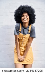 Portrait of joyful stylish African American hipster young woman wearing trendy yellow sundress with afro hair standing against white bricks wall background smiling looking at camera.