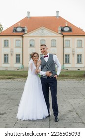 Portrait Of Joyful Wedding Couple Standing Outside Near Old Building In Summer. Young Gorgeous Woman Bride Hugging Young Man Groom, Embracing, Looking At Camera. Relationship, Love, Celebration.