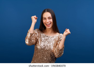  Portrait of joyful young attractive brunette woman in sparkling dress with goldent sequins, clenching her fists making winner's gesture, celebrating victory in online lottery, she hit the jackpot