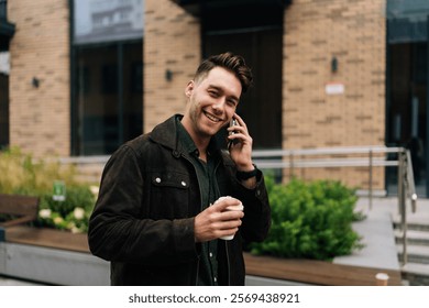 Portrait of joyful young man in casual attire standing waiting by modern building, talking on smartphone and holding cup of takeaway coffee, enjoying break alone. Concept of successful city lifestyle. - Powered by Shutterstock