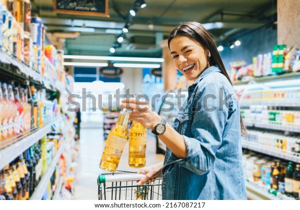 Portrait joyful young woman buying beer in liquor\
store holding two bottles in her hand winking cheerfully smiling\
looking at camera