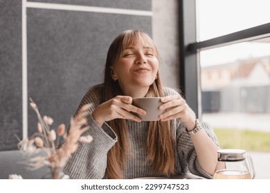 Portrait of joyful young woman enjoying a cup of tea and aroma at restaurant. Smiling pretty girl drinking hot tea in cafe. Excited blonde woman with bang wearing grey sweater and laugh, closed eyes. - Powered by Shutterstock