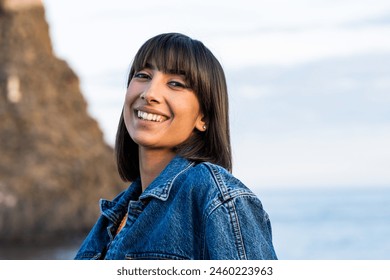 Portrait of a joyful young woman wearing a denim jacket, smiling brightly at the seaside with natural rock formations in the background. - Powered by Shutterstock