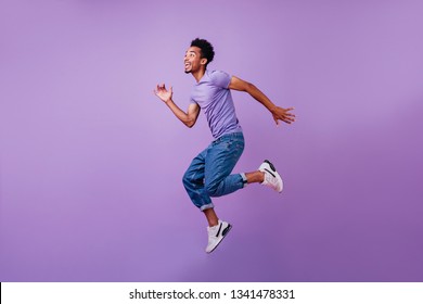 Portrait of jumping amazed guy in white sneakers. Indoor shot of dancing stylish male model in purple t-shirt.