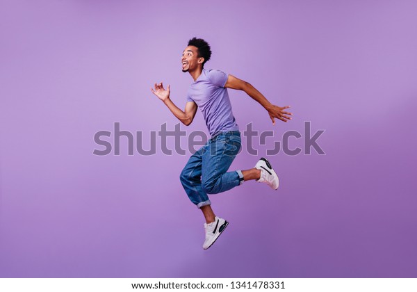 Portrait of jumping\
amazed guy in white sneakers. Indoor shot of dancing stylish male\
model in purple\
t-shirt.