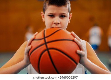 Portrait of a junior athlete standing at indoor basketball court with a ball in his hands and looking at the camera. Young basketball kid is posing with a basketball in his hands. Sporty kid with ball - Powered by Shutterstock