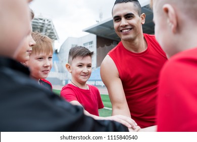 Portrait Of Junior Football Team Stacking Hands During Motivational Pep Talk Before Match In Outdoor Stadium, Focus On Handsome Coach, Copy Space