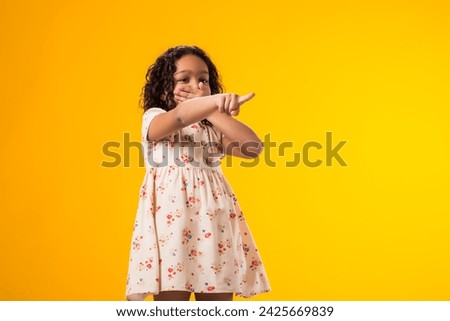 Portrait of kid girl mocking and teasing at someone showing finger at camera and closing her mouth over yellow background. Bulling concept