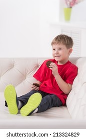 Portrait Of Kid Sitting In White Sofa. Little Boy Eating Sweets And Smiling