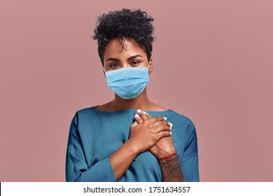 Portrait of kind hearted African American female paramedic wearing a respiratory mask from coronavirus disease, keeps hands on chest, shows her kindness and sympathy, on beige wall. COVID-19 epidemic - Shutterstock ID 1751634557