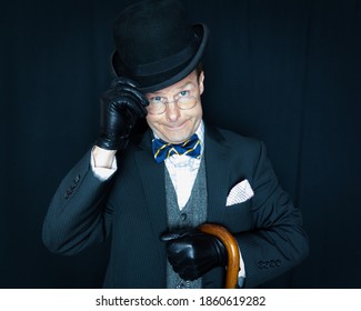 Portrait Of Kind Man In Dark Suit And Leather Gloves Doffing A Bowler Hat On Black Background. Stereotypical British Gentleman. Vintage Fashion. Retro Style. Polite And Courteous. Sharp Dressed Man.