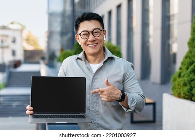 Portrait Of Korean Businessman Pointing At Laptop Computer With Blank Black Screen, Standing Outdoors, Mockup. People, Technologies And Remote Work Concept