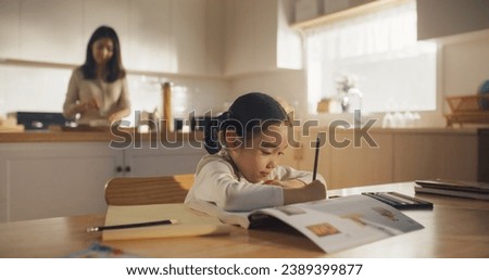 Portrait of a Korean Female Child Sitting at a Kitchen Table and Drawing While her Mother is Preparing Breakfast. Little Cute Girl Waiting for Nutritious Meal Before Going to School