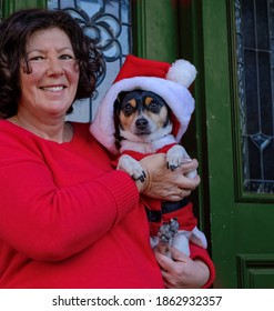 Portrait Of Lap Dog Dressed Like Santa Claus; Smiling Mature Female Owner In Red Sweater On The Side; Green Front Door In Background