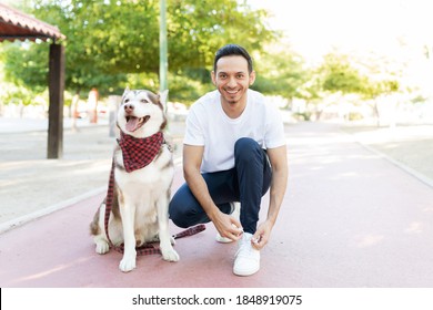 Portrait Of A Latin Adult Man Tying His Shoelaces Next To His Sitting Big Dog And Preparing For A Run In The Park 