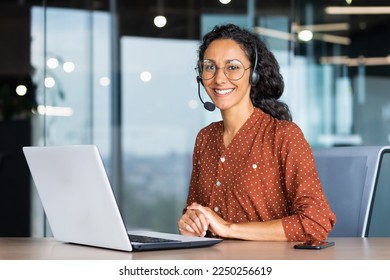 Portrait of Latin American business woman, office worker looking at camera and smiling, using headset and laptop for remote online communication, customer support tech call center worker. - Powered by Shutterstock