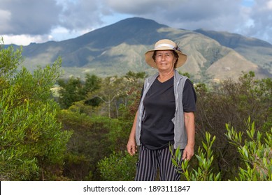 Portrait Of Latin Old Farmer Woman On The Field With Shrubbery And Mountains In Background