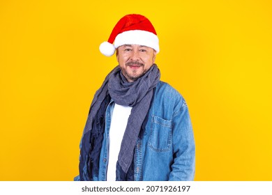 Portrait Of Latin Senior Man With Santa Hat For Christmas Party On Yellow Background In Mexico Latin America