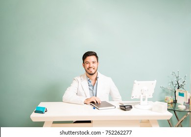 Portrait Of A Latin Young Therapist With A Lab Coat Sitting In The Front Desk Of A Health And Spa Clinic And Smiling