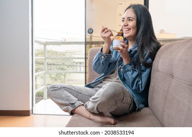 Portrait Of A Latina Woman Smiling While Eating Homemade Cashew Butter While Sitting On A Couch At Home