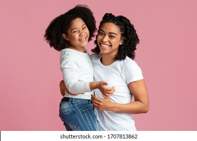 Portrait of laughing black mother and daughter cuddling and looking at camera, having fun together, isolated on pink background with copy space